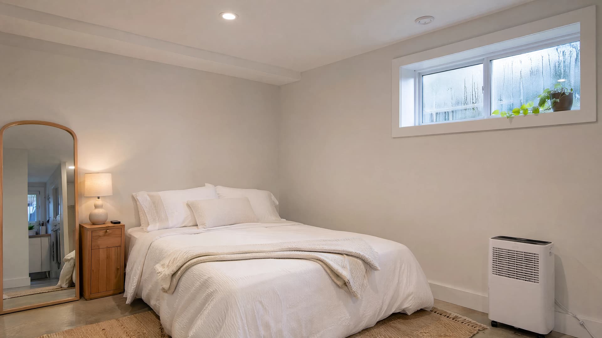 Compact dehumidifier running in a basement suite bedroom with rain condensation on the small high window